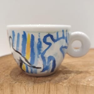 Op de afbeelding: Een witte keramische espressokop met een handgeschilderd abstract ontwerp met verticale blauwe en gele penseelstreken, geaccentueerd door gedurfde zwarte contouren van gestileerde figuren. De kop heeft een dik, rond handvat en een glanzende afwerking.