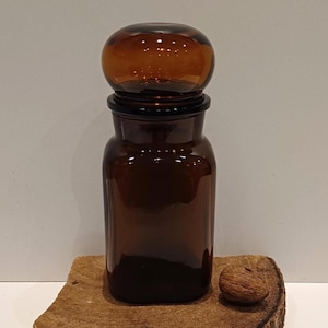 Könnte beinhalten: Ein antikes Apothekerglas aus braunem Glas mit einem runden Verschluss. Das Glas ist zylindrisch mit breitem Boden und schmalem Hals. Der Verschluss ist eine abgerundete Kugel. Das Glas steht auf einem Holzstück.