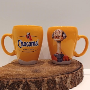 May include: Two yellow ceramic mugs. One mug features the Chocomel logo in blue and white. The other mug has a whimsical illustration of a figure with a long neck and a detailed face. Both mugs have a curved handle.