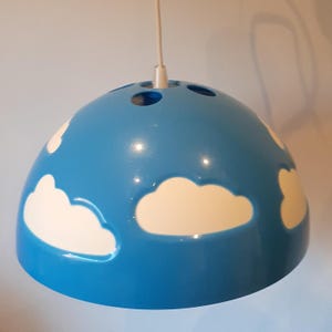 Może przedstawiać: Niebieska lampa wisząca z białymi wzorami chmur. Oprawa ma zaokrąglony kształt z kilkoma małymi otworami u góry. Chmury są namalowane na powierzchni oprawy.