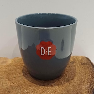 May include: A dark grey ceramic pot with a glossy finish. A red, flower-shaped sticker with the white letters "D.E" is affixed to the front. The pot is sitting on a wooden surface.