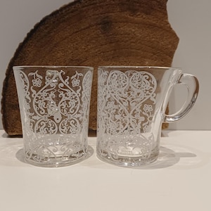 May include: Two clear glass mugs with a decorative white floral pattern. One mug has a handle, and the other is a tumbler. The mugs are labeled "Douwe Egberts" at the bottom. The background features a piece of brown wood.