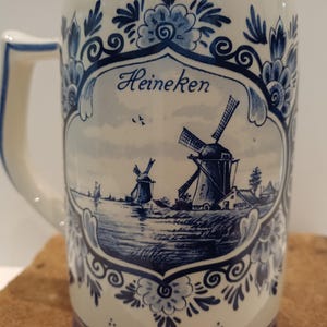 May include: A ceramic beer mug featuring a blue and white floral border and a central illustration of two windmills near a body of water. The word Heineken is printed in blue script above the windmills. The mug has a white handle on the left side.