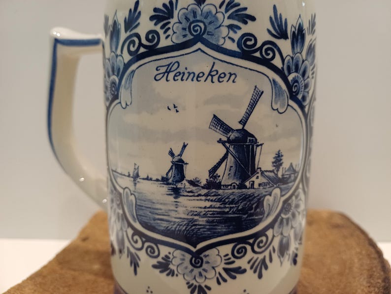 May include: A ceramic beer mug featuring a blue and white floral border and a central illustration of two windmills near a body of water. The word Heineken is printed in blue script above the windmills. The mug has a white handle on the left side.