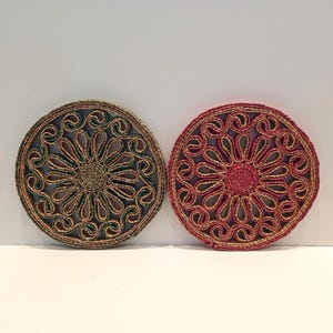Könnte beinhalten: Zwei runde Untersetzer mit aufwendigen Blumenmustern. Einer ist hauptsächlich grün und braun, der andere rot und beige. Beide haben eine gewebte Textur und eine dunkelblaue Rückseite. Schützen Oberflächen vor Hitze und Feuchtigkeit.