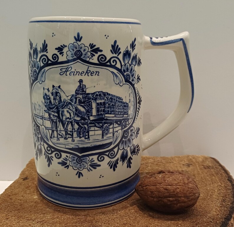 May include: A cream-colored ceramic beer mug featuring a blue transferware design. The central illustration depicts a horse-drawn carriage with the text Heineken above it. The mug is decorated with ornate blue floral patterns and a blue base rim.