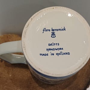 May include: The underside of a white ceramic mug featuring blue text that reads flora keramiek, a small symbol, DELFTS, HANDWORK, and MADE IN HOLLAND. The mug has a curved handle extending from the side and a smooth, circular base rim.