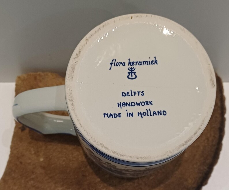 May include: The underside of a white ceramic mug featuring blue text that reads flora keramiek, a small symbol, DELFTS, HANDWORK, and MADE IN HOLLAND. The mug has a curved handle extending from the side and a smooth, circular base rim.