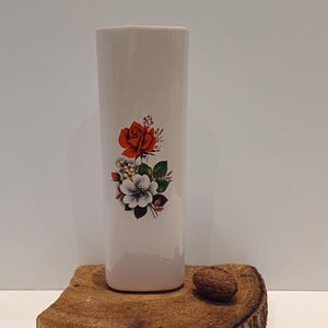 May include: White ceramic vase with a floral design featuring a red rose and white flowers. The vase has a rectangular shape and is approximately 6 inches tall.