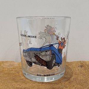 May include: A clear glass tumbler featuring a cartoon scene with a cowboy in a blue car, shooting a gun. The glass has the text "SUSKE & WISKE" and "BOB & BOBETTE" printed on it. The tumbler is set on a wooden surface.