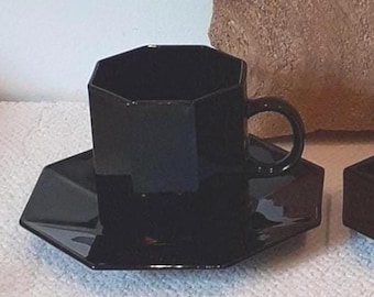 May include: A black, geometric teacup and saucer set. The cup and saucer have a faceted design, with a glossy finish. The cup has a rounded handle. The set is displayed on a white surface.