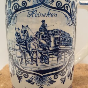 May include: A cream-colored ceramic beer mug featuring a blue transferware design. The central illustration depicts a horse-drawn carriage carrying barrels, with the text Heineken above. The mug is decorated with ornate blue floral patterns.