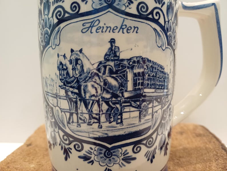 May include: A cream-colored ceramic beer mug featuring a blue transferware design. The central illustration depicts a horse-drawn carriage carrying barrels, with the text Heineken above. The mug is decorated with ornate blue floral patterns.