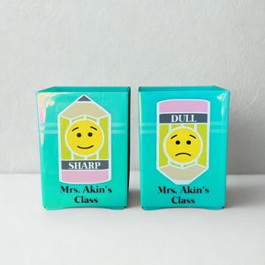 May include: Two teal square pencil holders featuring pencil-shaped decals. The left holder displays a smiling face with the text SHARP and Mrs. Akin's Class. The right holder displays a frowning face with the text DULL and Mrs. Akin's Class.