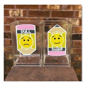 May include: Two clear rectangular acrylic pencil holders featuring vinyl decals. The left holder displays a yellow pencil with a sad face and the text DULL. The right holder shows a yellow pencil with a happy face and the text SHARP. Both containers are labeled Mrs. Hailey's Class.