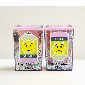 May include: Two clear rectangular pencil holders filled with small multicolored foam beads. Each holder features a pencil graphic with a yellow smiley face, the text Mrs. Amador's Class, and either the word SHARP or the word DULL printed on them.