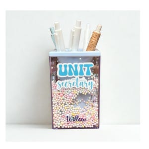 May include: A rectangular clear acrylic pen holder filled with small multicolored foam beads. The front features the text "UNIT secretary" in blue and white, with the name "Willow" printed in dark purple script at the bottom, holding several pens.