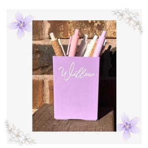 May include: A light purple rectangular pen holder featuring the name Willow in white script lettering. The container is filled with several pens, including pink, white, and cork-textured writing instruments, standing upright inside the holder.