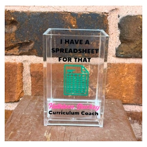 May include: A clear acrylic rectangular pencil holder featuring the black text I HAVE A SPREADSHEET FOR THAT, a teal graphic of a spreadsheet icon, the pink name Kathleen Stuckey, and the black text Curriculum Coach printed on the front surface.