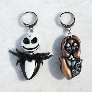 Puede incluir: Un par de pendientes con Jack Skellington y Sally de Pesadilla antes de Navidad. El pendiente de Jack es blanco y negro, mientras que el de Sally es marrón, negro y dorado. Ambos pendientes tienen aros plateados.