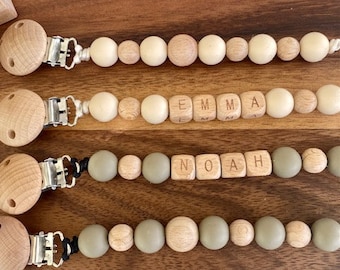 Pode incluir: Três colares de dentição de madeira com clipes. O colar superior tem contas de cor creme e contas de letras de madeira que soletram "EMMA". O colar do meio tem contas cinzas e contas de letras de madeira que soletram "NOAH". O colar inferior tem contas creme e cinzas.