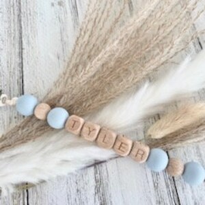 Pode incluir: Um colar de nome para beb&ecirc; personalizado com o nome "TYLER" escrito em blocos de madeira. O colar &eacute; feito com contas de silicone azul claro e contas de madeira.