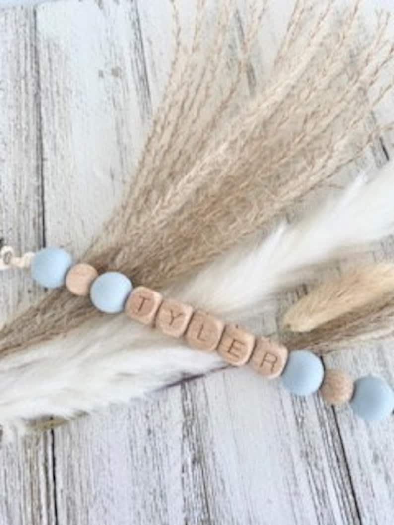 Pode incluir: Um colar de nome para beb&ecirc; personalizado com o nome "TYLER" escrito em blocos de madeira. O colar &eacute; feito com contas de silicone azul claro e contas de madeira.
