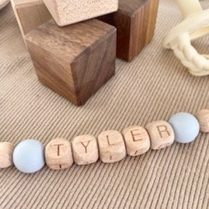 Pode incluir: Um colar de nome para beb&ecirc; de madeira com o nome "TYLER" escrito em contas de madeira individuais. As contas s&atilde;o intercaladas com contas de silicone azul claro.