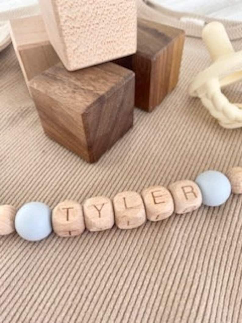 Pode incluir: Um colar de nome para beb&ecirc; de madeira com o nome "TYLER" escrito em contas de madeira individuais. As contas s&atilde;o intercaladas com contas de silicone azul claro.
