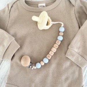 Pode incluir: Um prendedor de chupeta para beb&eacute; em madeira natural e azul claro com uma conta de madeira e uma conta de silicone. O prendedor &eacute; preso a uma chupeta com um anel de silicone amarelo. O prendedor tem o nome "LYDIA" escrito em contas de madeira.