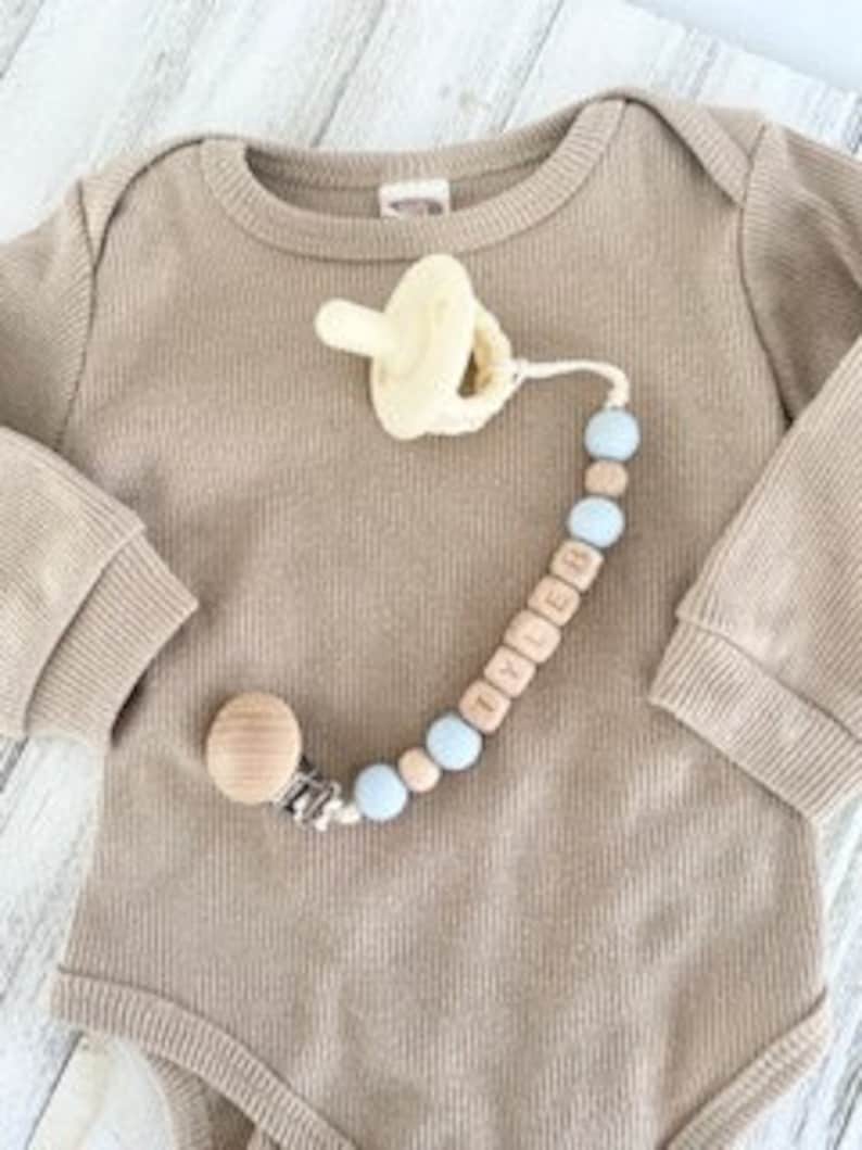 Pode incluir: Um prendedor de chupeta para beb&eacute; em madeira natural e azul claro com uma conta de madeira e uma conta de silicone. O prendedor &eacute; preso a uma chupeta com um anel de silicone amarelo. O prendedor tem o nome "LYDIA" escrito em contas de madeira.