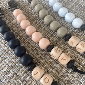 May include: A set of five silicone and wooden baby teethers with personalized wooden beads. The teethers are in various colors, including black, white, beige, and brown. The wooden beads are engraved with the letters "J.D", "M.K", and "K.S".