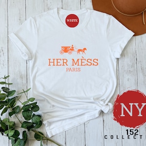Puede incluir: Camiseta blanca con un gr&aacute;fico naranja de un carruaje tirado por un caballo y el texto "HER M&Eacute;S PARIS". La camiseta est&aacute; sobre un fondo de madera blanca con un c&iacute;rculo rojo con el texto "NY 152 COLLECTIVE" en blanco.