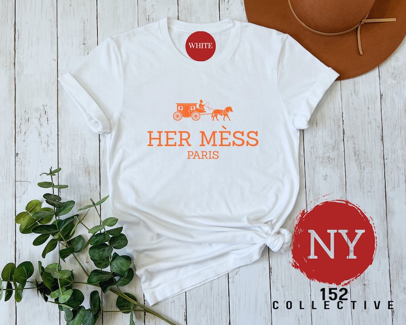 Puede incluir: Camiseta blanca con un gr&aacute;fico naranja de un carruaje tirado por un caballo y el texto "HER M&Eacute;S PARIS". La camiseta est&aacute; sobre un fondo de madera blanca con un c&iacute;rculo rojo con el texto "NY 152 COLLECTIVE" en blanco.