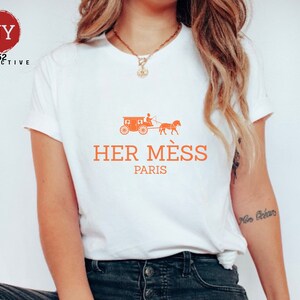 Puede incluir: Camiseta blanca con un gr&aacute;fico naranja de un carruaje tirado por un caballo y el texto "HER M&Eacute;S PARIS".