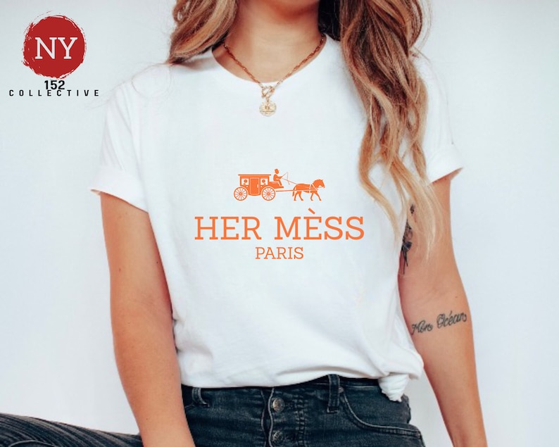 Puede incluir: Camiseta blanca con un gr&aacute;fico naranja de un carruaje tirado por un caballo y el texto "HER M&Eacute;S PARIS".