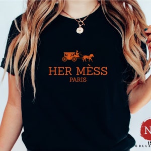 Puede incluir: Camiseta negra con un gr&aacute;fico naranja de un carruaje tirado por un caballo y el texto "HER MESS PARIS".