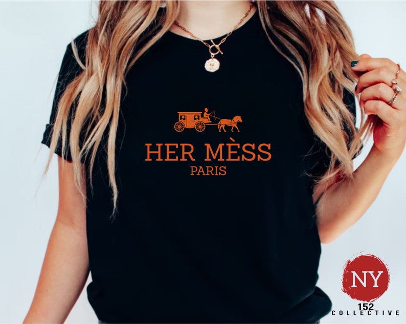 Puede incluir: Camiseta negra con un gr&aacute;fico naranja de un carruaje tirado por un caballo y el texto "HER MESS PARIS".