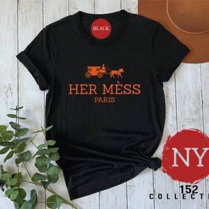 Puede incluir: Camiseta negra con un gr&aacute;fico naranja de un carruaje tirado por un caballo y el texto "HER M&Eacute;S PARIS".