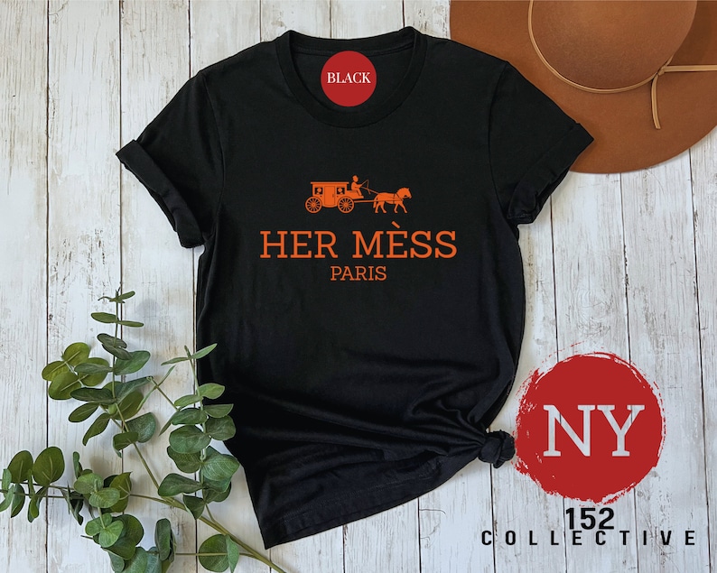 Puede incluir: Camiseta negra con un gr&aacute;fico naranja de un carruaje tirado por un caballo y el texto "HER M&Eacute;S PARIS".