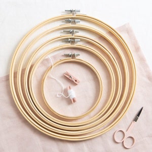 May include: A set of six wooden embroidery hoops of varying sizes, nested together. The hoops are on a pink fabric surface with a small ball of white yarn and two wooden clothespins. A pair of pink handled scissors is in the lower right corner.