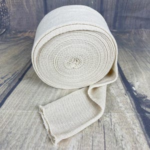 May include: A roll of natural beige fabric bandage, rolled up and tied with a knot. The fabric is woven and has a slightly textured surface.
