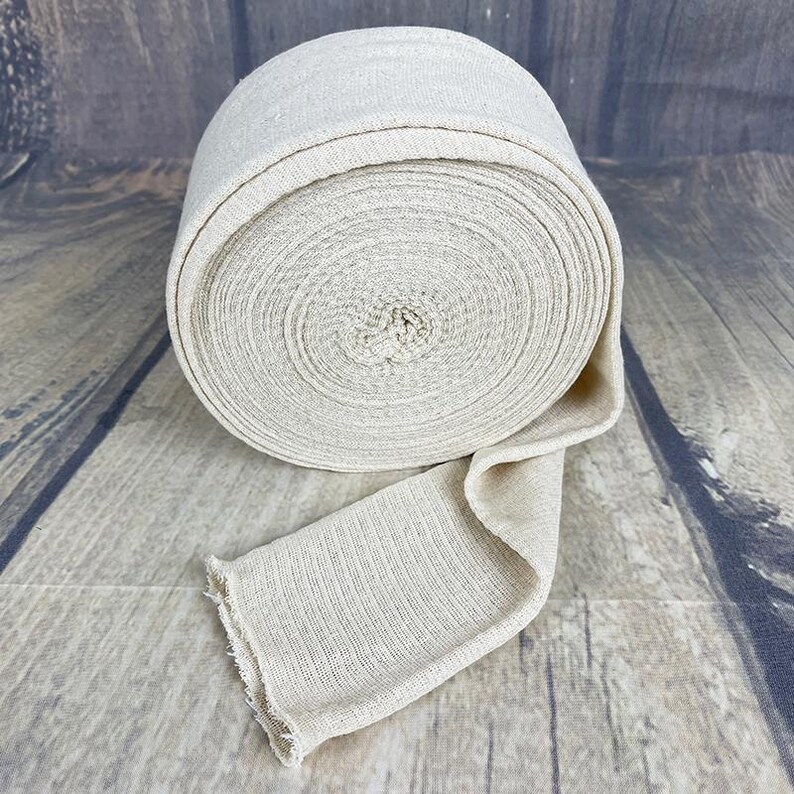 May include: A roll of natural beige fabric bandage, rolled up and tied with a knot. The fabric is woven and has a slightly textured surface.