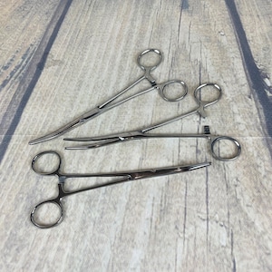 Puede incluir: Tres hemostáticos de acero inoxidable, también conocidos como pinzas, se colocan sobre una superficie de madera. Los hemostáticos son instrumentos quirúrgicos que se utilizan para sujetar vasos sanguíneos o tejidos.