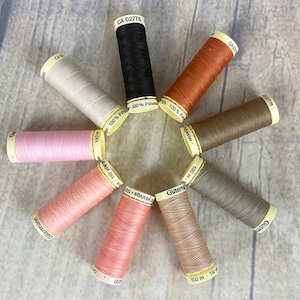 May include: A collection of ten spools of thread in various colors, including black, beige, pink, orange, brown, and gray. The spools are arranged in a circle on a wooden surface. Each spool has a label with the brand name "Gütermann" and the text "100 m 110 yds/vgs".