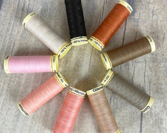 May include: A collection of ten spools of thread in various colors, including black, beige, pink, orange, brown, and gray. The spools are arranged in a circle on a wooden surface. Each spool has a label with the brand name "Gütermann" and the text "100 m 110 yds/vgs".