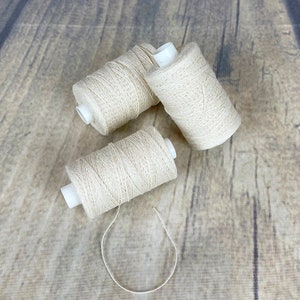 May include: Three spools of natural white cotton thread on a wooden surface.