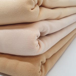 Op de afbeelding: Een stapel gevouwen stof in verschillende tinten beige en bruin. De stof lijkt een zacht gebreid materiaal te zijn, met zichtbare ribbels. De vouwen creëren een gelaagd effect.