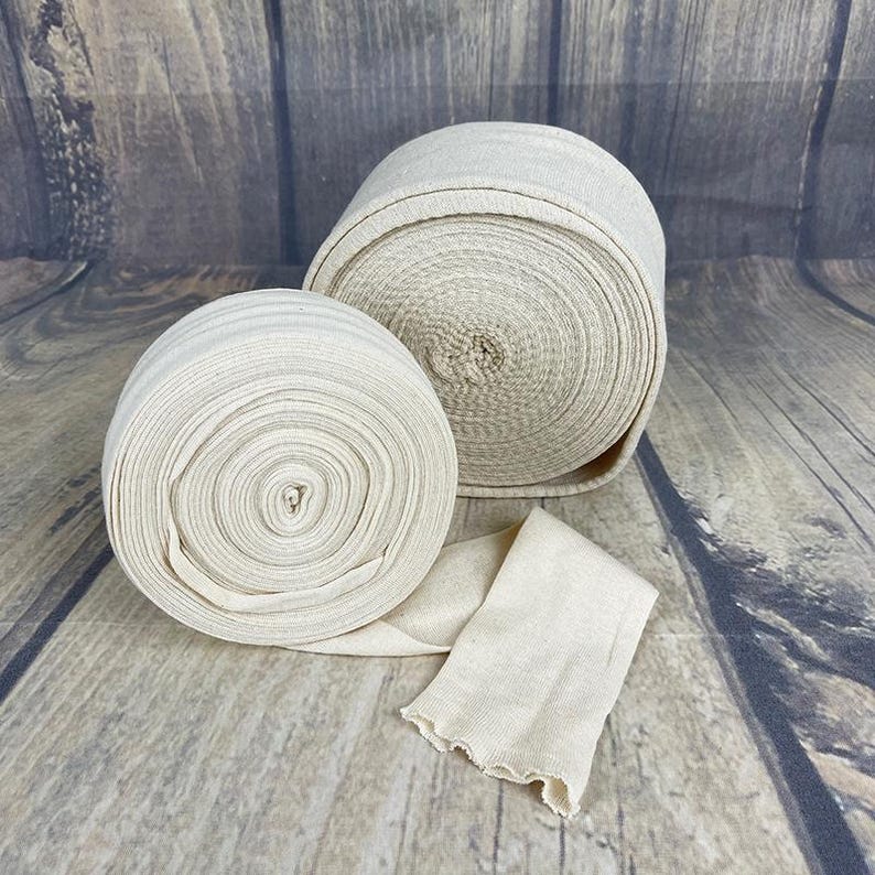 May include: Two rolls of natural colored fabric bandage, one larger and one smaller, with a separate piece of fabric bandage lying on a wooden surface.