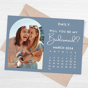 以下が含まれることがあります： 2人の女性が笑顔で写っている写真が特徴の、青いブライズメイドの提案カード。「EMILY, WILL YOU BE MY Bridesmaid?」のテキストと、3月2024年のカレンダーで15日を強調。封筒も写っています。